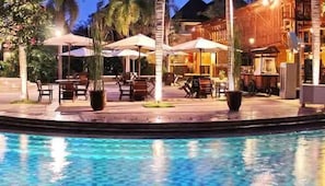 A poolside area with outdoor seating and umbrellas, surrounded by palm trees and a building with a warm facade. - LE DIAN HOTEL & COTTAGES (Serang)