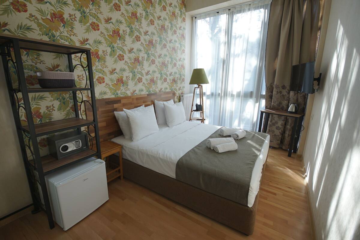 A neatly arranged bedroom with a bed, a small refrigerator, a shelf with a basket, a lamp, and a television on a stand.