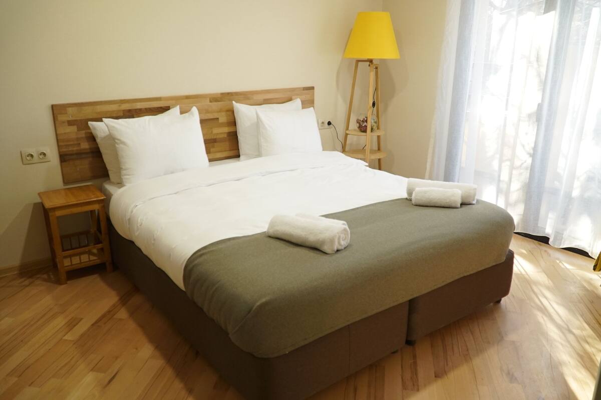 A neatly made bed with white linens and two towels, a wooden headboard, a bedside table with a lamp, and a window with sheer curtains.