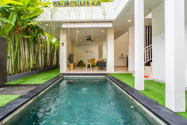 A modern indoor pool area with a clear view of the interior, including a seating area with a 'LOVE' sign, a ceiling fan, and a staircase leading to another level.