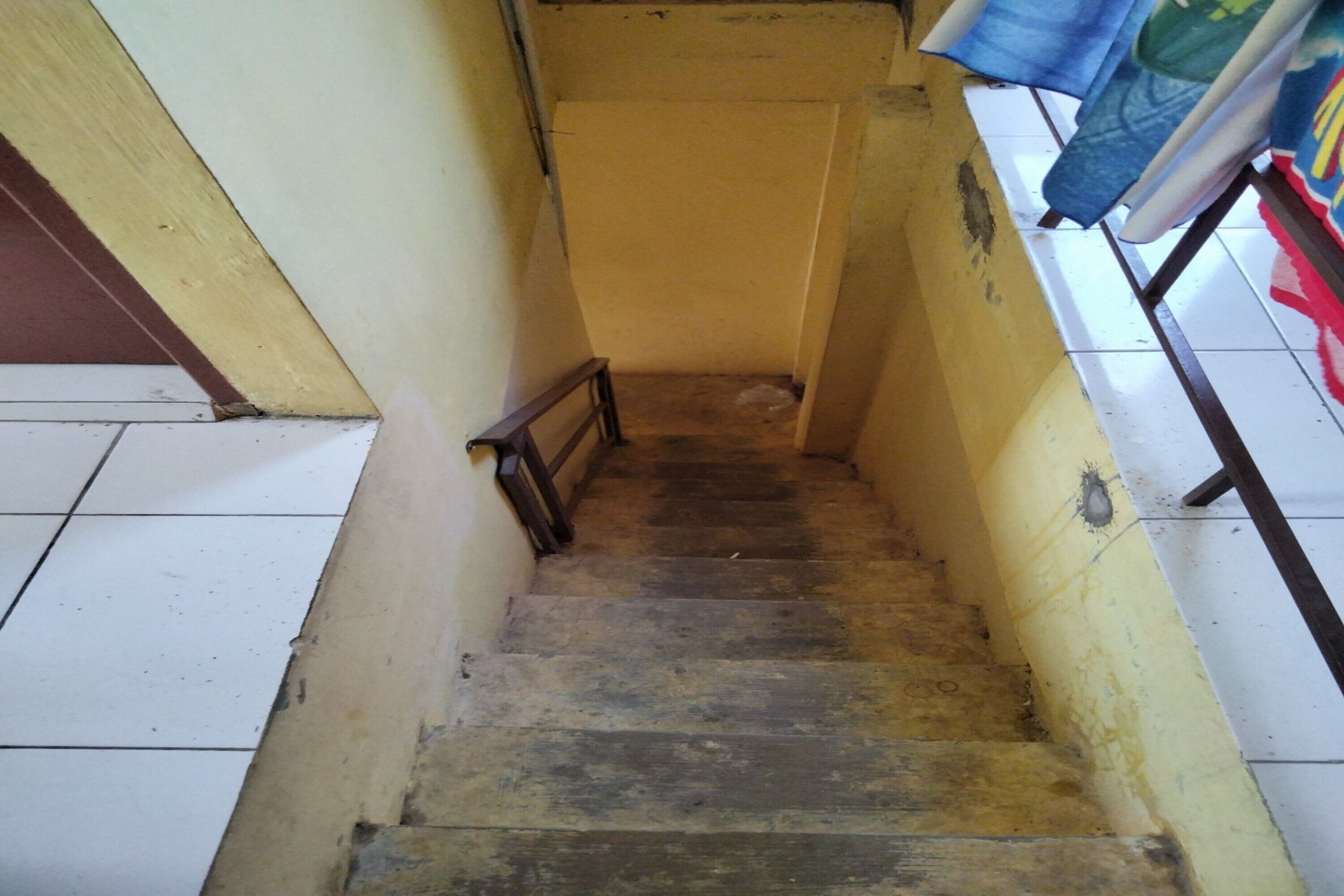A narrow staircase with wooden steps and a metal handrail, leading to a landing with a yellow wall and a tiled floor.