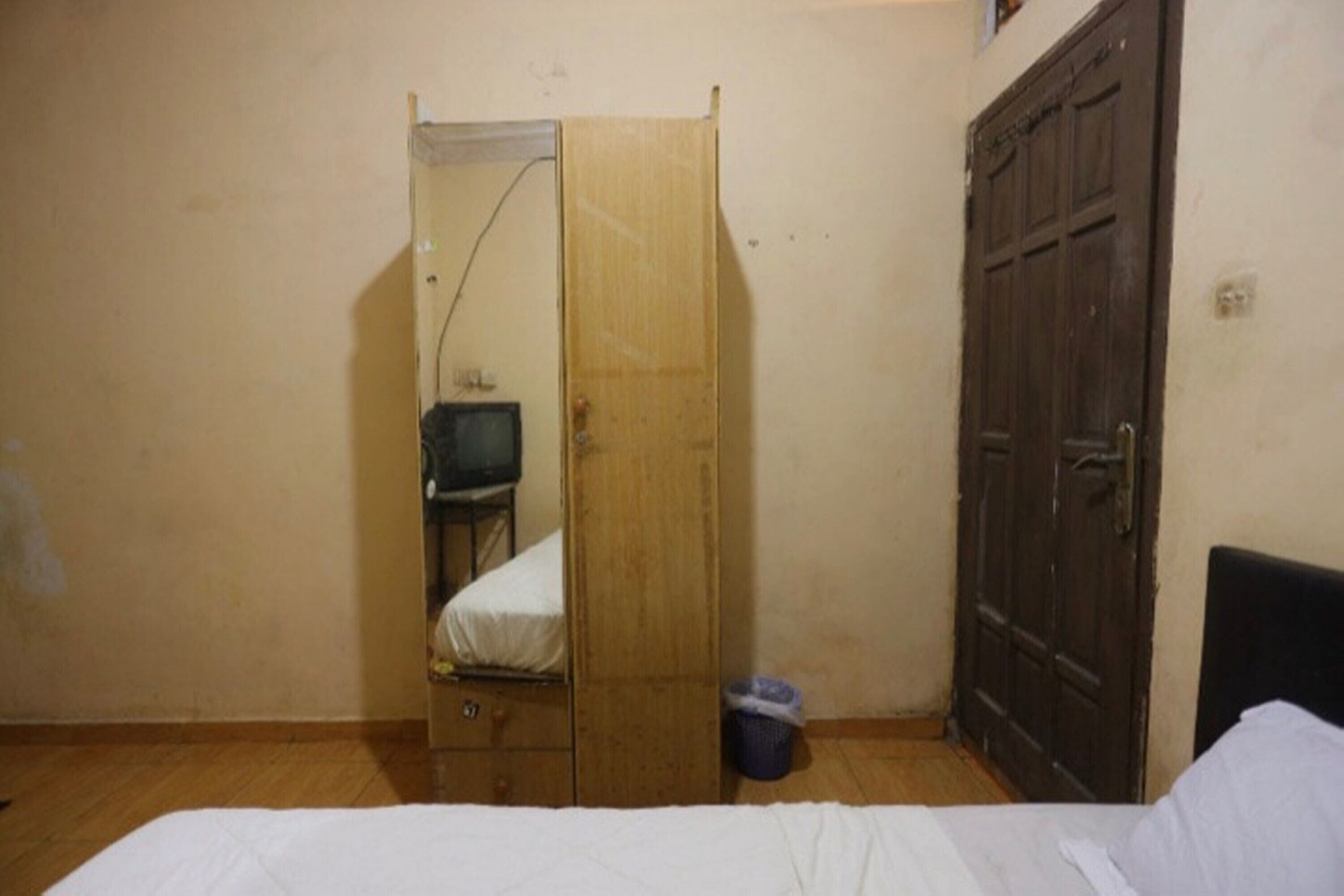A small, sparsely furnished room with a wooden wardrobe, a single bed, and a closed door.