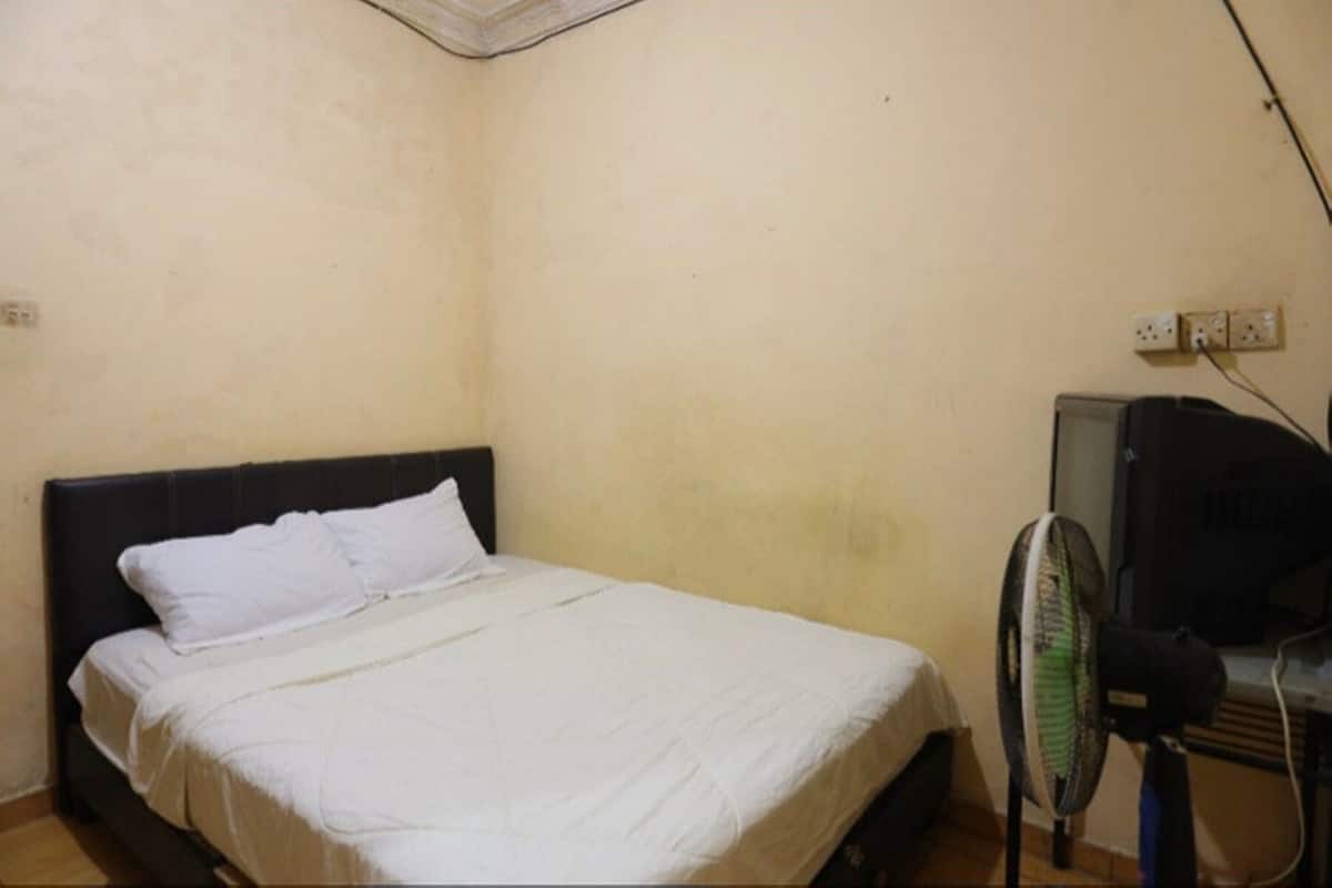 Una habitación pequeña con cama individual, pared sencilla, enchufe y ventilador.