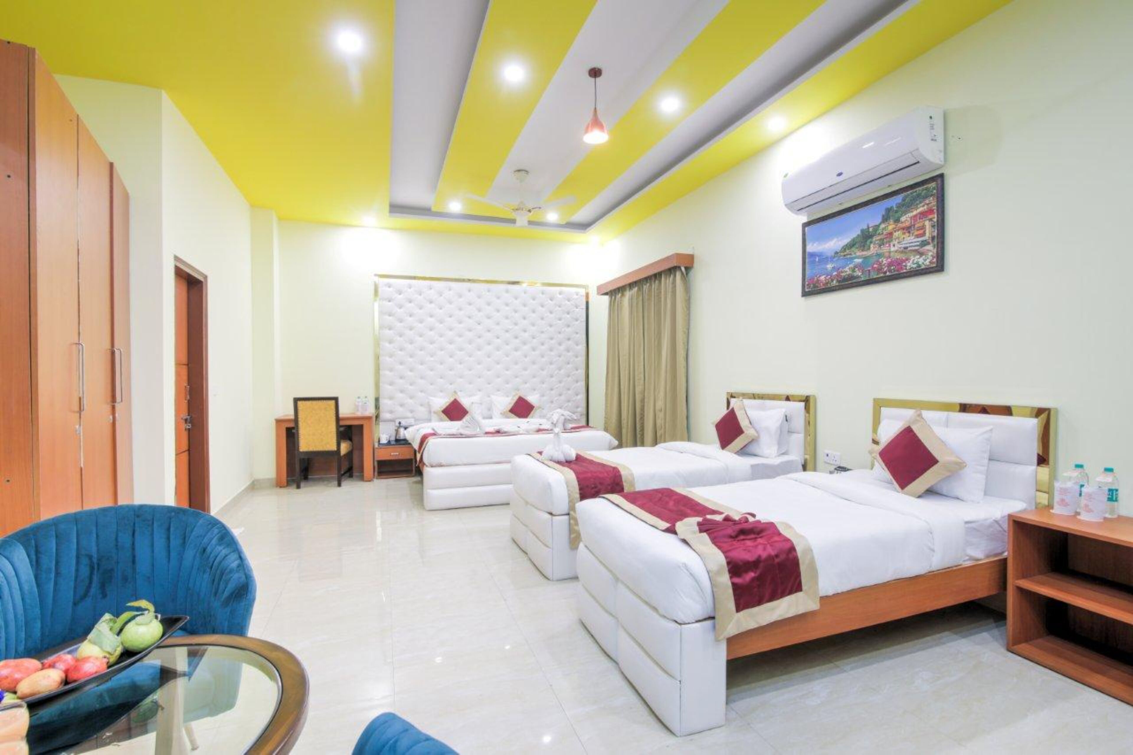 A hotel room with two beds, a dining table with fruit, a blue chair, and a TV on the wall.