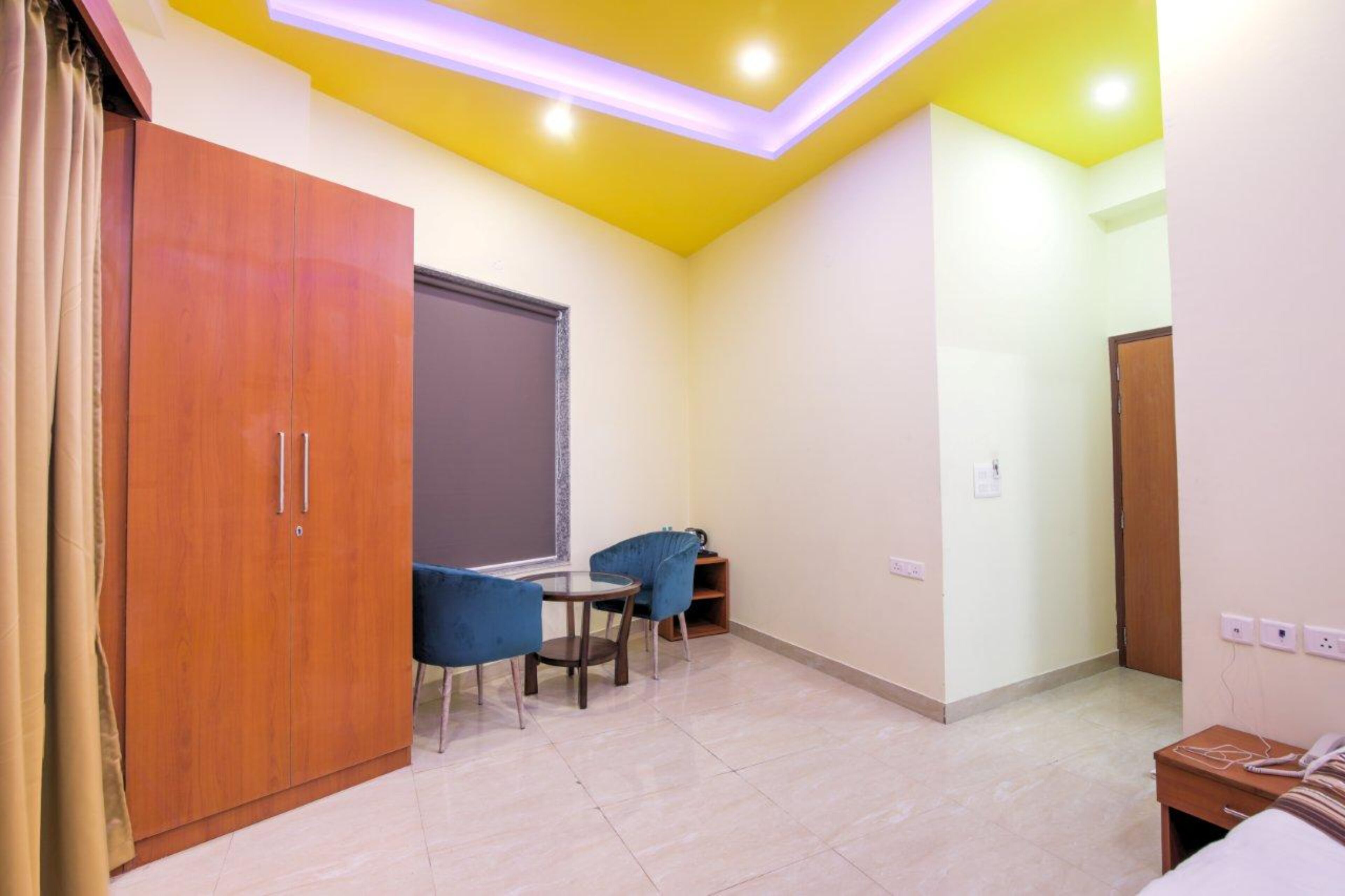 A room with a wooden wardrobe, a small round table with two chairs, a window with a blind, and a door.