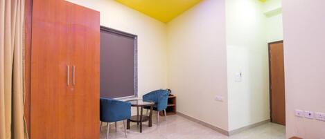A room with a wooden wardrobe, a small round table with two chairs, a window with a blind, and a door.