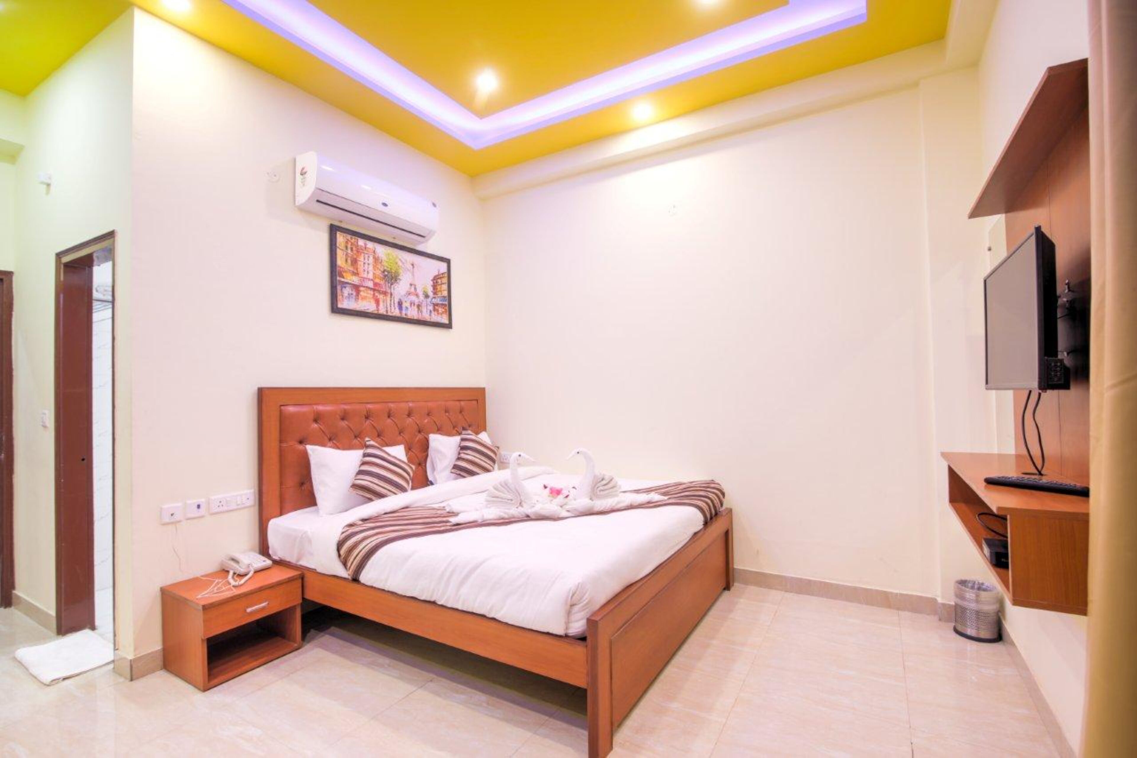 A hotel room with a bed, bedside table, air conditioning unit, wall-mounted TV, and a painting.