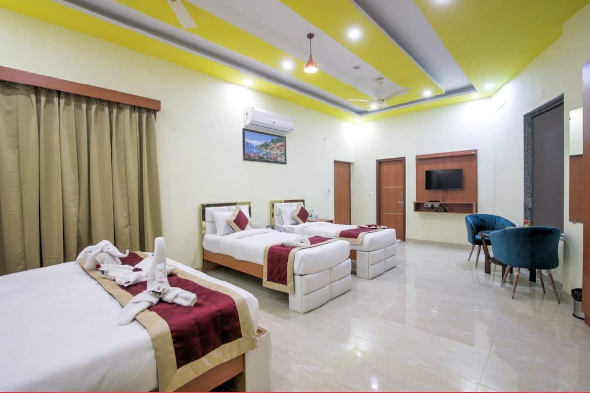 Quarto de hotel com três camas, área de refeições com duas cadeiras e uma televisão fixada na parede.