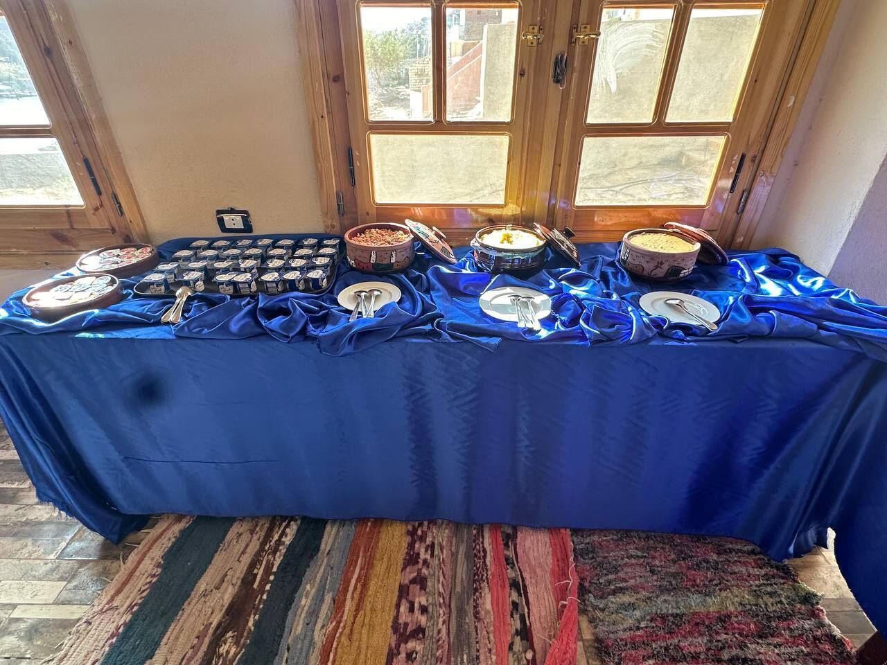 Una mesa de buffet con diversos alimentos y utensilios, sobre un mantel azul, con una alfombra estampada y puertas de madera al fondo.