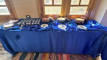 Una mesa de buffet con diversos alimentos y utensilios, sobre un mantel azul, con una alfombra estampada y puertas de madera al fondo.