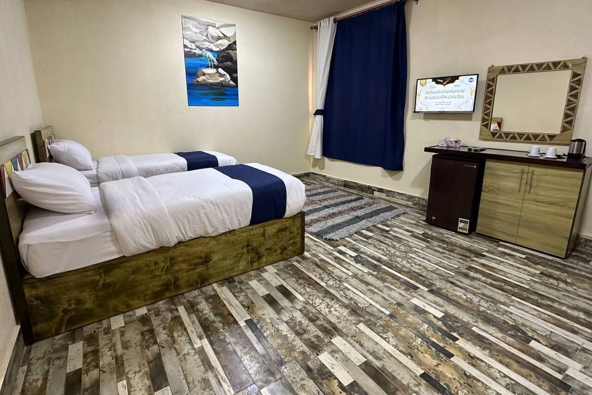 A bedroom with a bed, a desk, a small refrigerator, a mirror, and a painting on the wall.