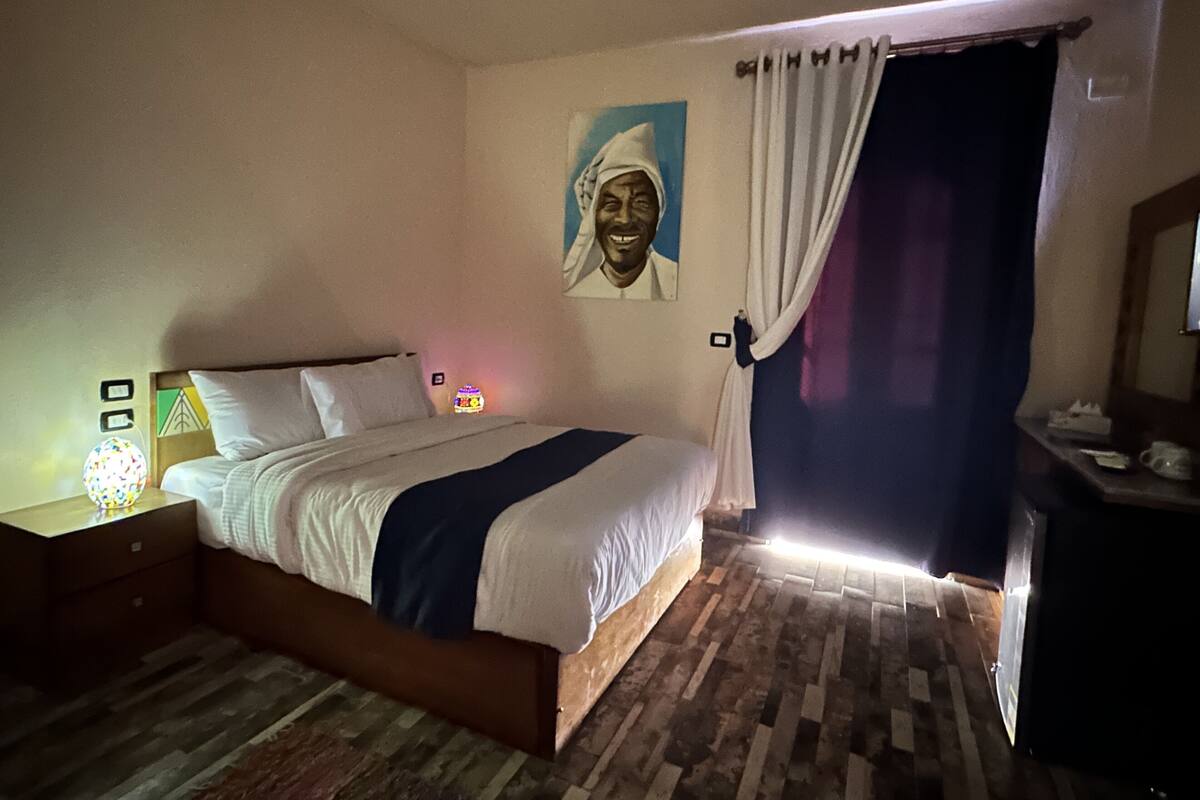 A bedroom with a bed, a nightstand, a wall-mounted lamp, a painting, and a curtain.
