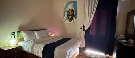 Quarto com cama, mesa de cabeceira, luminária na parede, um quadro e cortina.