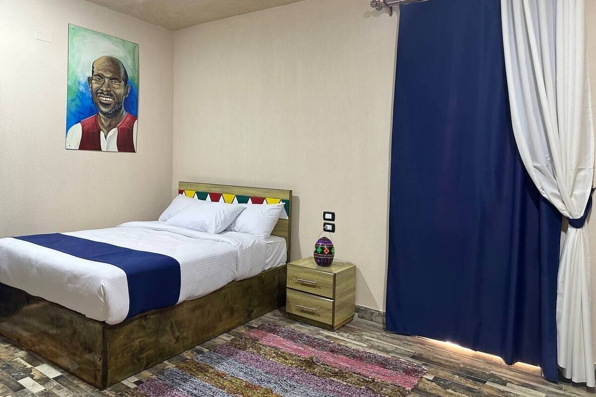 A bedroom with a bed, a nightstand, a painting, and a curtain.