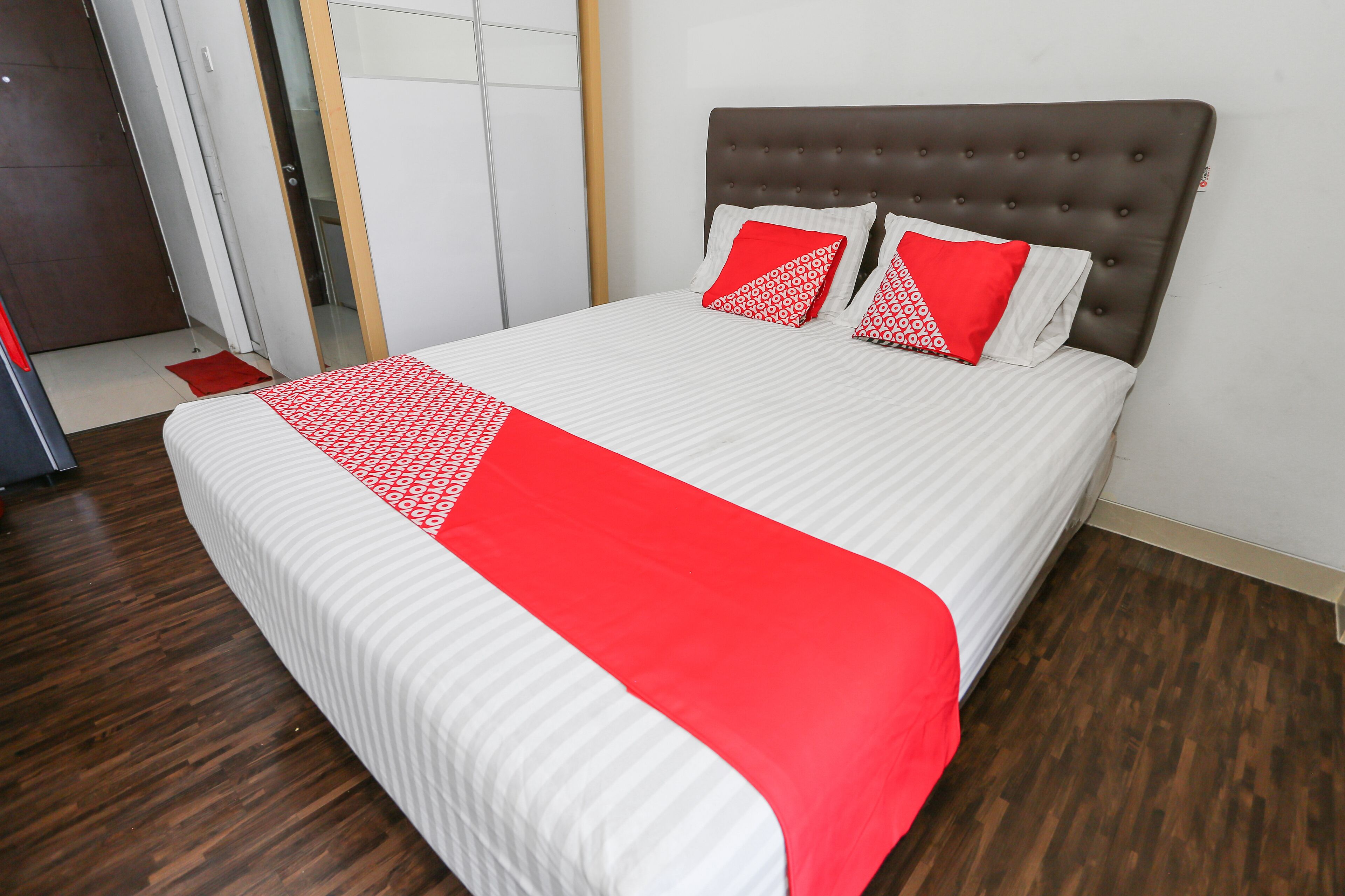 Een net opgemaakt bed met een wit-rode sprei, een bruin gestoffeerd hoofdbord en twee rode kussens met een wit patroon.