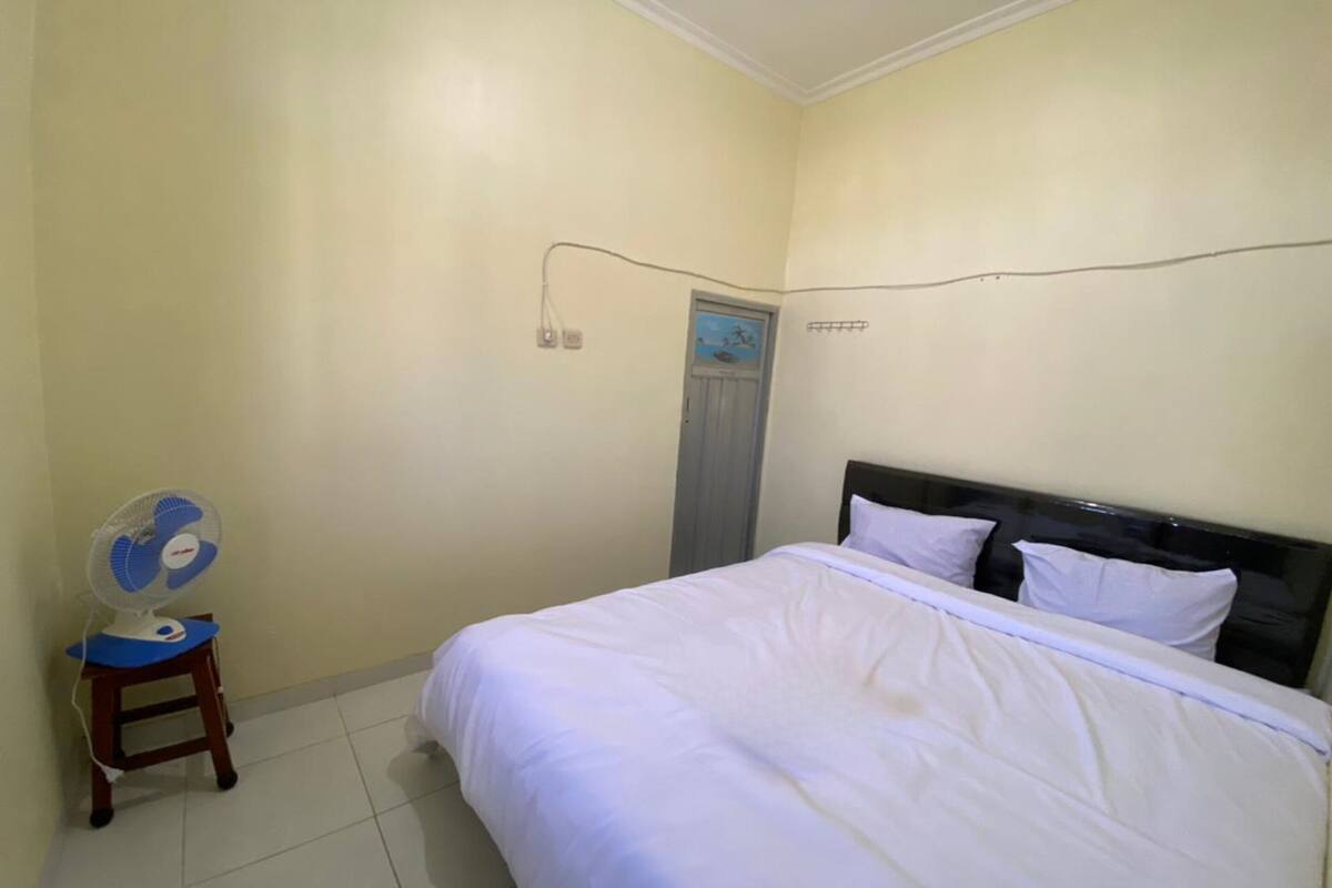 A bedroom with a bed, white bedding, a wooden nightstand, a blue and white fan, and a wall socket.