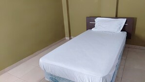 Una cama individual con colchón y almohada blancos en una habitación con paredes lisas y suelo de baldosas.