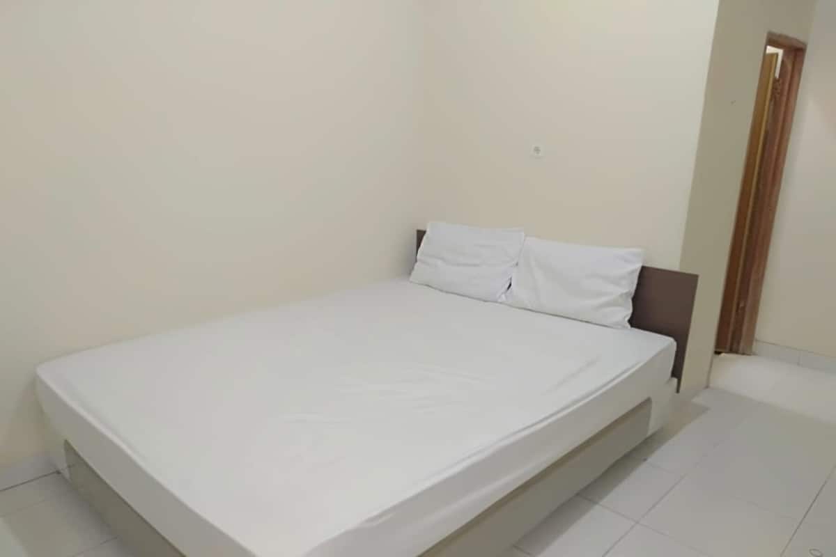 A single-bed room with a white mattress and headboard, a wooden door, and white tiled flooring.