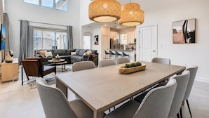 A modern living room with a dining area, large windows, and a kitchen in the background.
