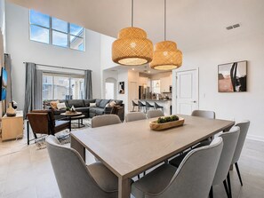 A modern living room with a dining area, large windows, and a kitchen in the background.