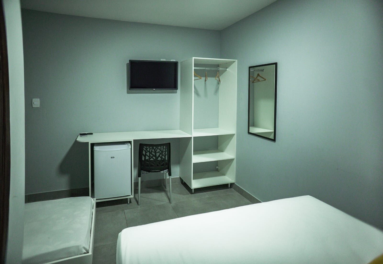 A hotel room with a bed, a desk with a chair, a small refrigerator, a wardrobe, and a television mounted on the wall.