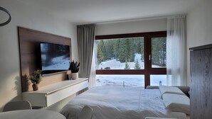 A hotel room with a bed, a TV mounted on the wall, a desk with a plant, and a view of a snowy landscape through large windows.