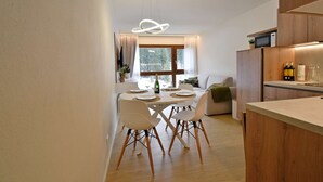 A modern kitchen with wooden cabinets, a dining area with white chairs, and a large window with curtains.