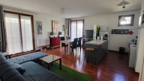 Een woonkamer met een bank, een eethoek met stoelen, een keuken met een koelkast en een klein schiereiland.