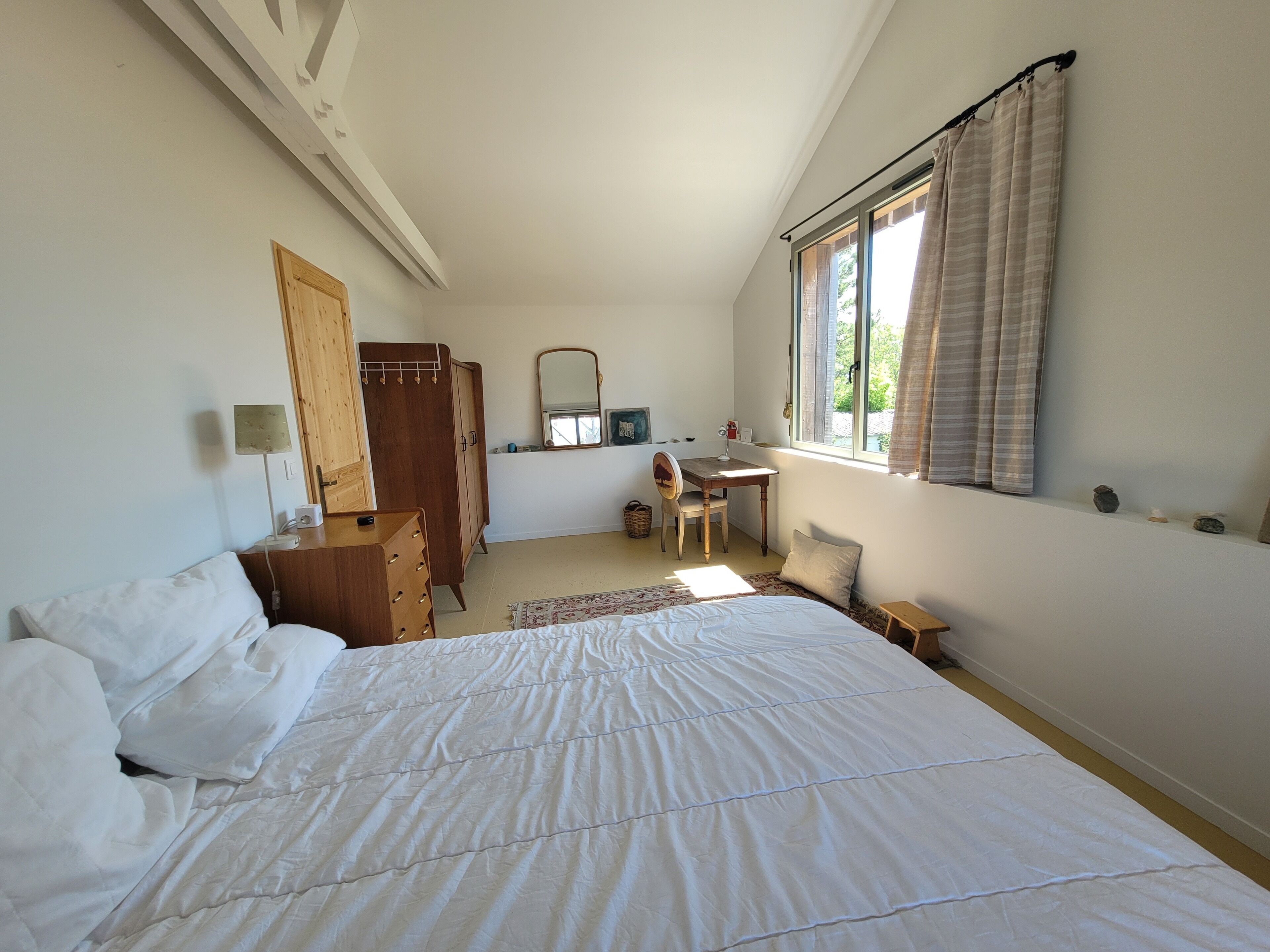 Une chambre avec un grand lit, une armoire en bois, un bureau avec une chaise, une fenêtre avec des rideaux et un miroir sur le mur.