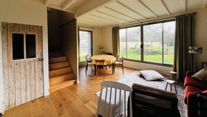Amplia sala de estar con escalera de madera, una mesa de comedor con sillas, un sofá y ventanas grandes que ofrecen vistas al exterior.
