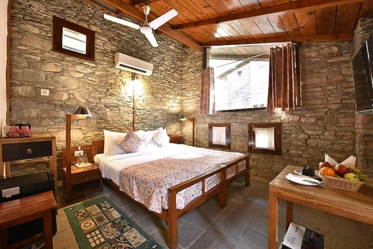 A rustic bedroom with a stone wall, wooden ceiling, a bed with a floral bedspread, a nightstand, a table with fruit, and a window with curtains.