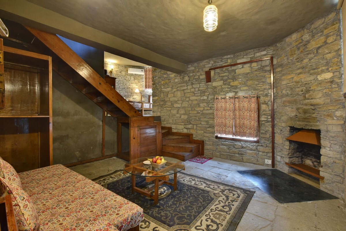 A rustic interior with a stone wall, wooden staircase, a glass table with a fruit bowl, and a patterned sofa.