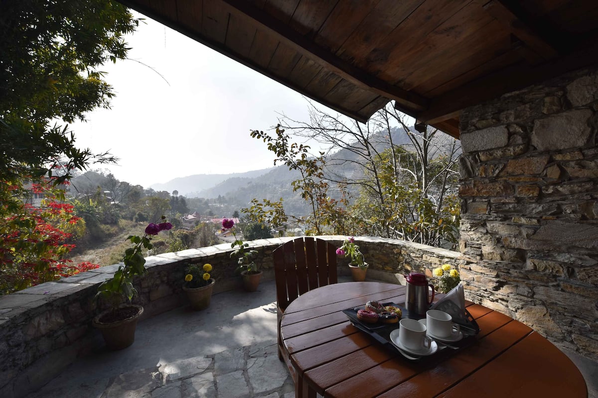 A wooden table with a tray of food and drinks, surrounded by potted plants, overlooking a mountainous landscape.