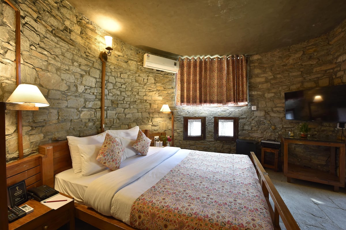 A bedroom with a stone wall, a wooden bed, a flat-screen TV, and a small table with a lamp.