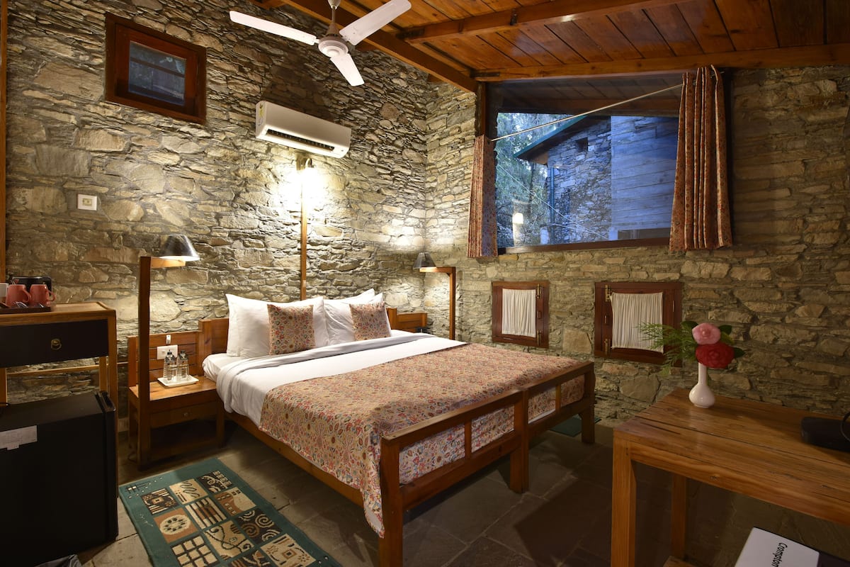 A bedroom with a stone wall, wooden ceiling, a bed with a floral bedspread, a nightstand with a lamp, a wooden table with a vase, and a window with curtains.