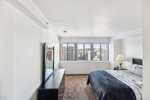 Un dormitorio con cama, cómoda, ventana con persianas y vista a la ciudad.