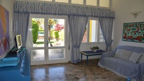 A room with a stone-tiled floor, a blue sofa, a small table with a vase, and a door leading to a garden. - Bright Villa in Caorle With Private Garden (Caorle)