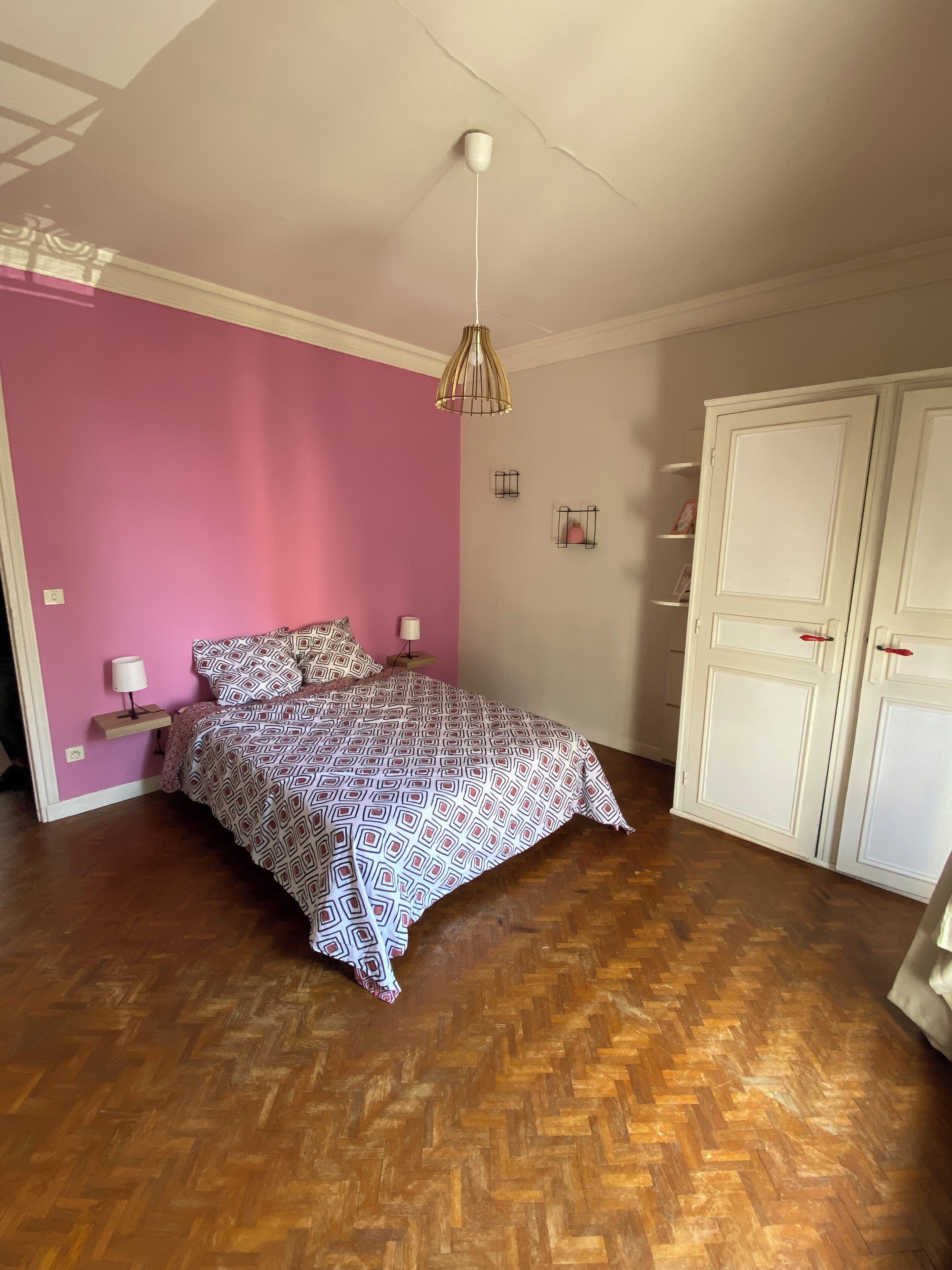 A bedroom with a patterned bedspread, a bedside lamp, a hanging light fixture, and a wardrobe.