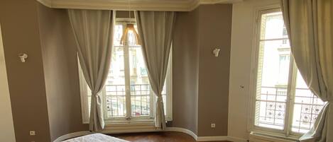 A spacious bedroom with a large bed, wooden parquet flooring, and two windows with curtains.