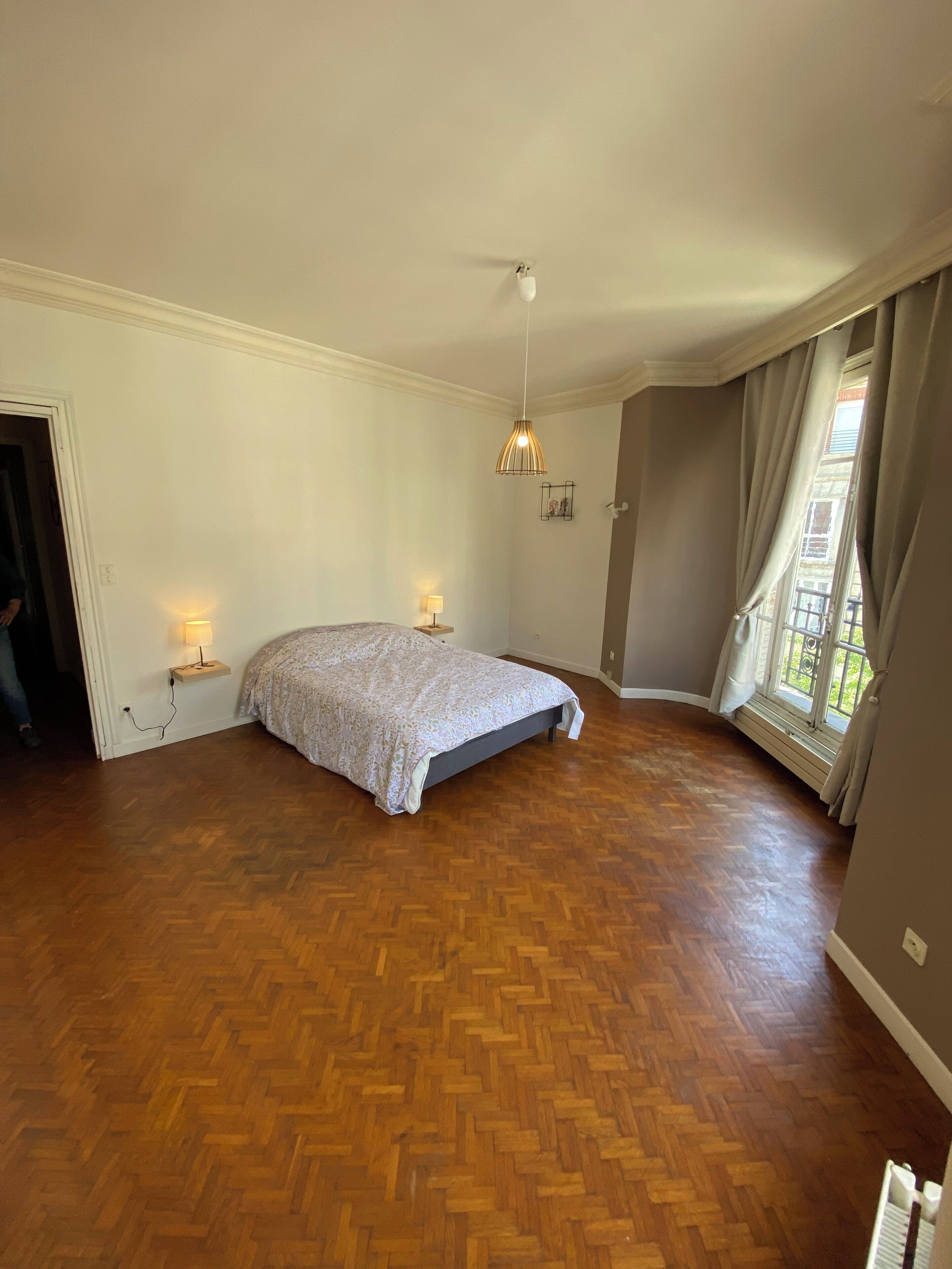 A spacious room with a large bed, wooden parquet flooring, a hanging light fixture, and a window with curtains.