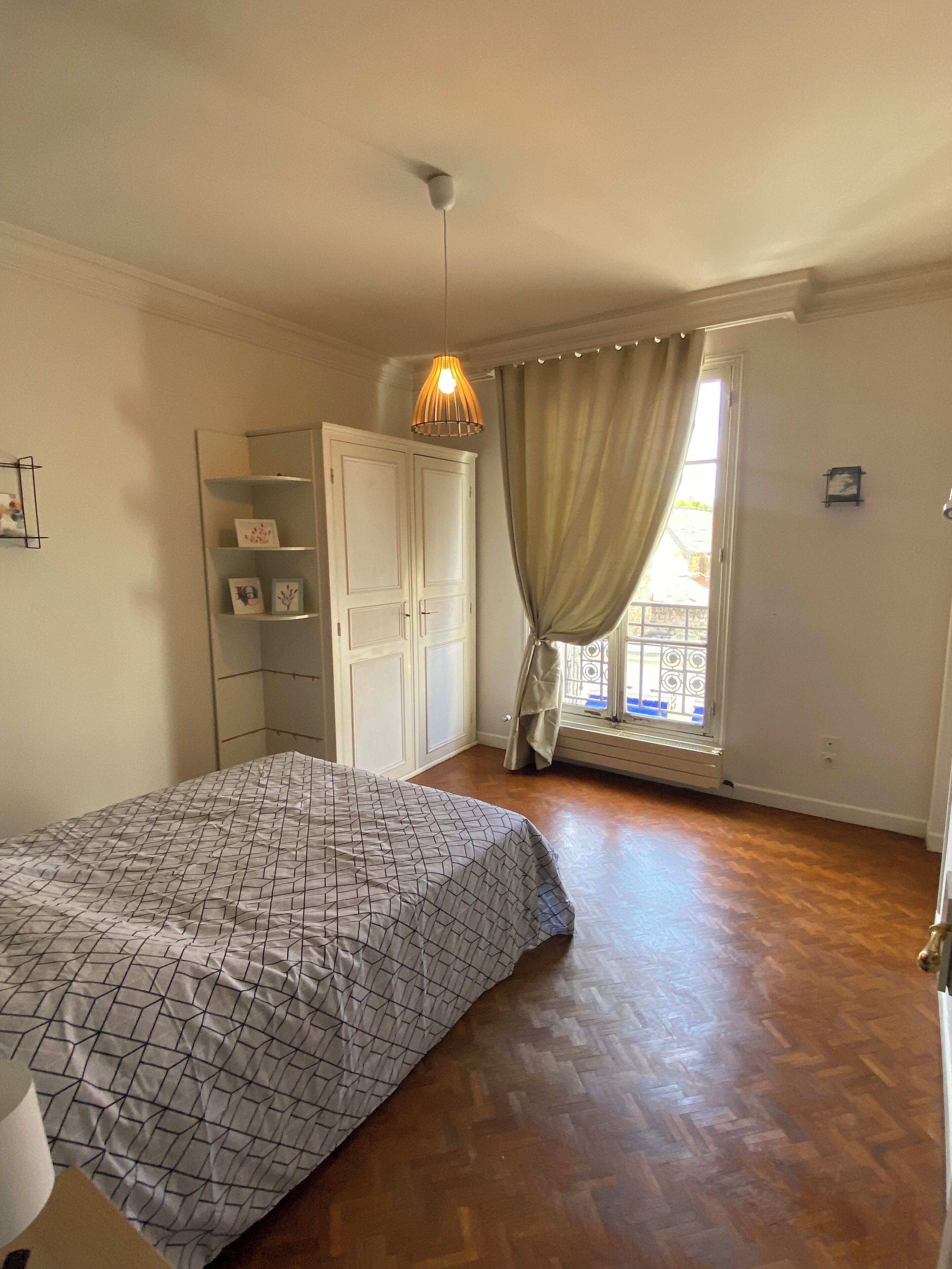 Un dormitorio con cama, armario, lámpara colgante y ventana con cortinas.