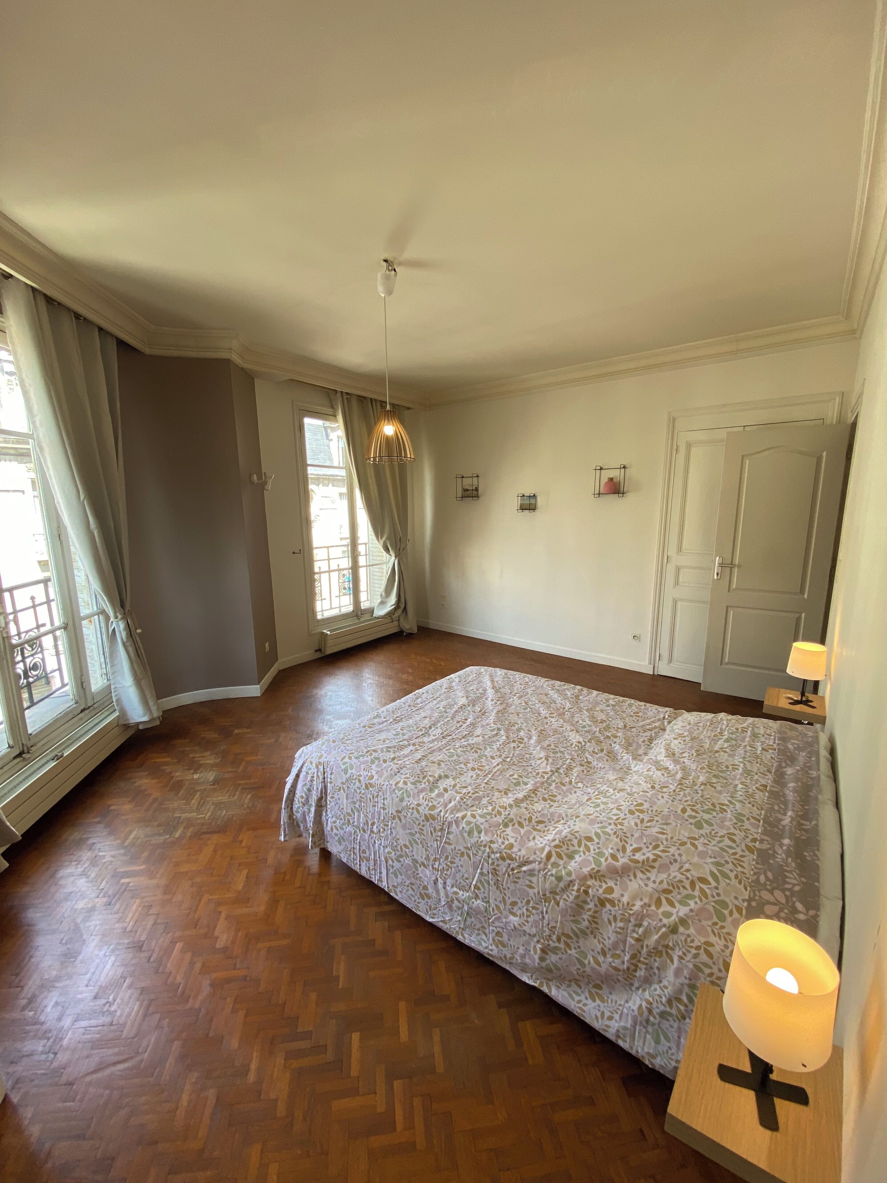 A bedroom with a large bed, wooden flooring, a hanging light fixture, and a door with a handle.