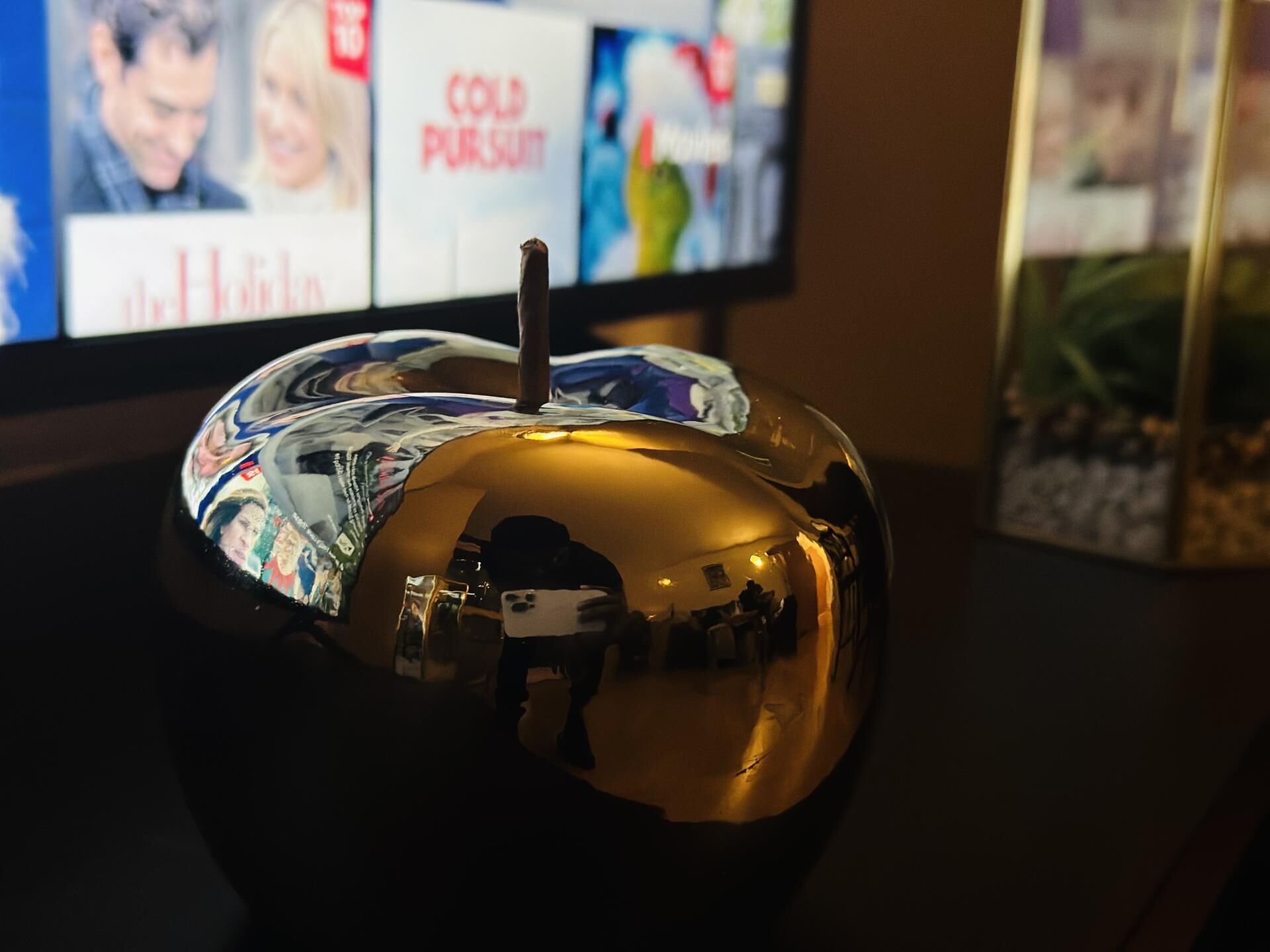 Un objeto esférico con superficie reflectante, posiblemente decorativo, sobre una superficie oscura con un televisor mostrando varias imágenes al fondo.
