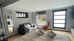 Een moderne slaapkamer met een groot bed, een bank, een bijzettafeltje en een stoel. Er is een raam met uitzicht op bomen en een deur.