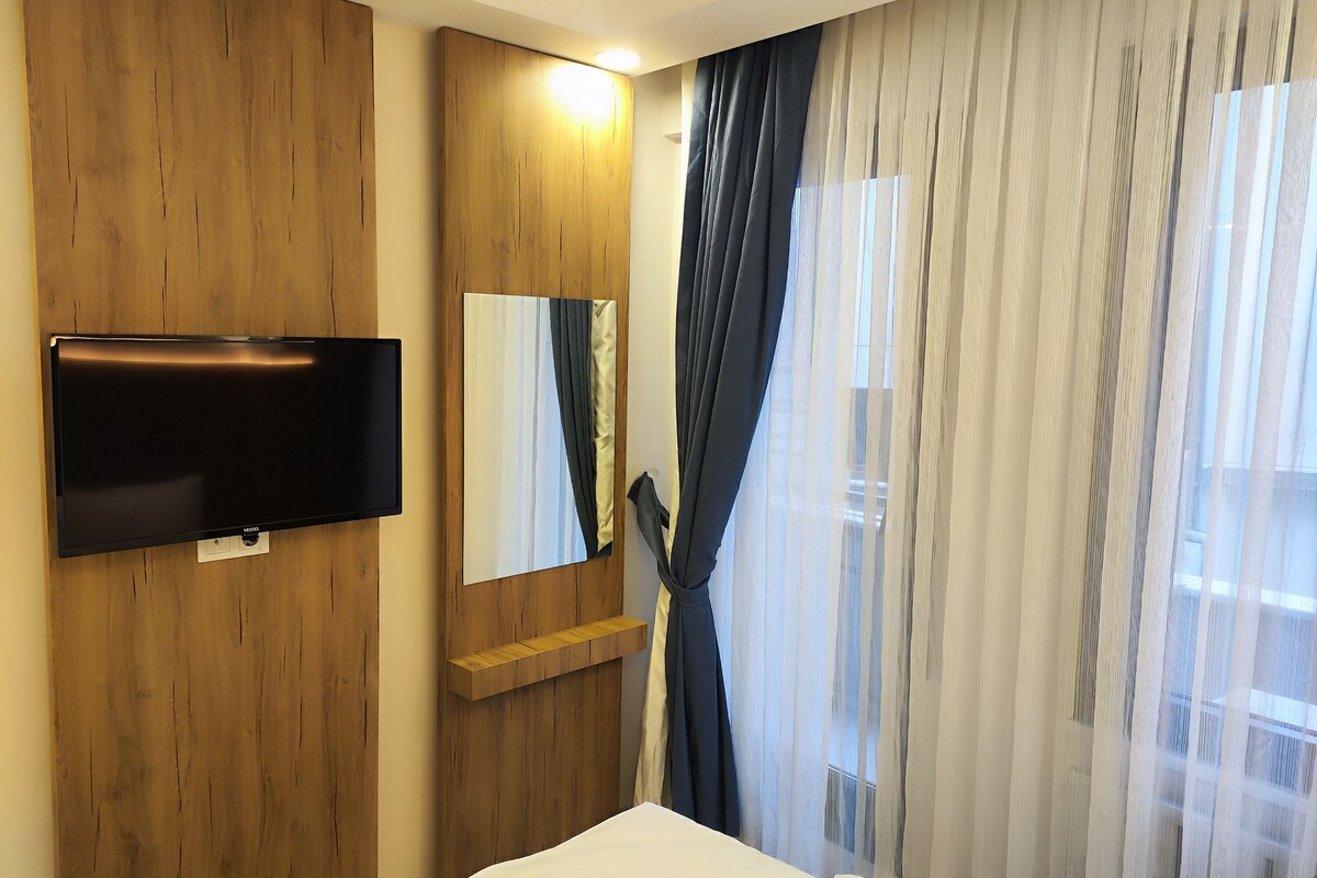 A hotel room with a flat-screen TV mounted on a wooden panel, a mirror, and a window with sheer curtains.