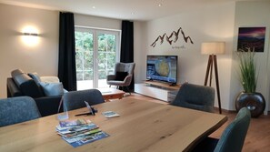 A modern living room with a wooden dining table, grey sofas, a TV, and a large window with curtains. - Fewo "zur Nagelfluh", Oberstaufen/steibis (Oberstaufen)