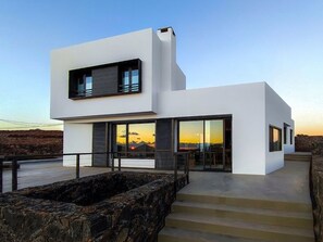 Une maison moderne à deux étages, dotée de grandes fenêtres, d’un mur de soutènement en pierre et d’un escalier menant à l’entrée.
