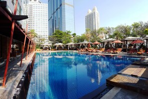 A swimming pool with lounge chairs and umbrellas, surrounded by tall buildings and trees.