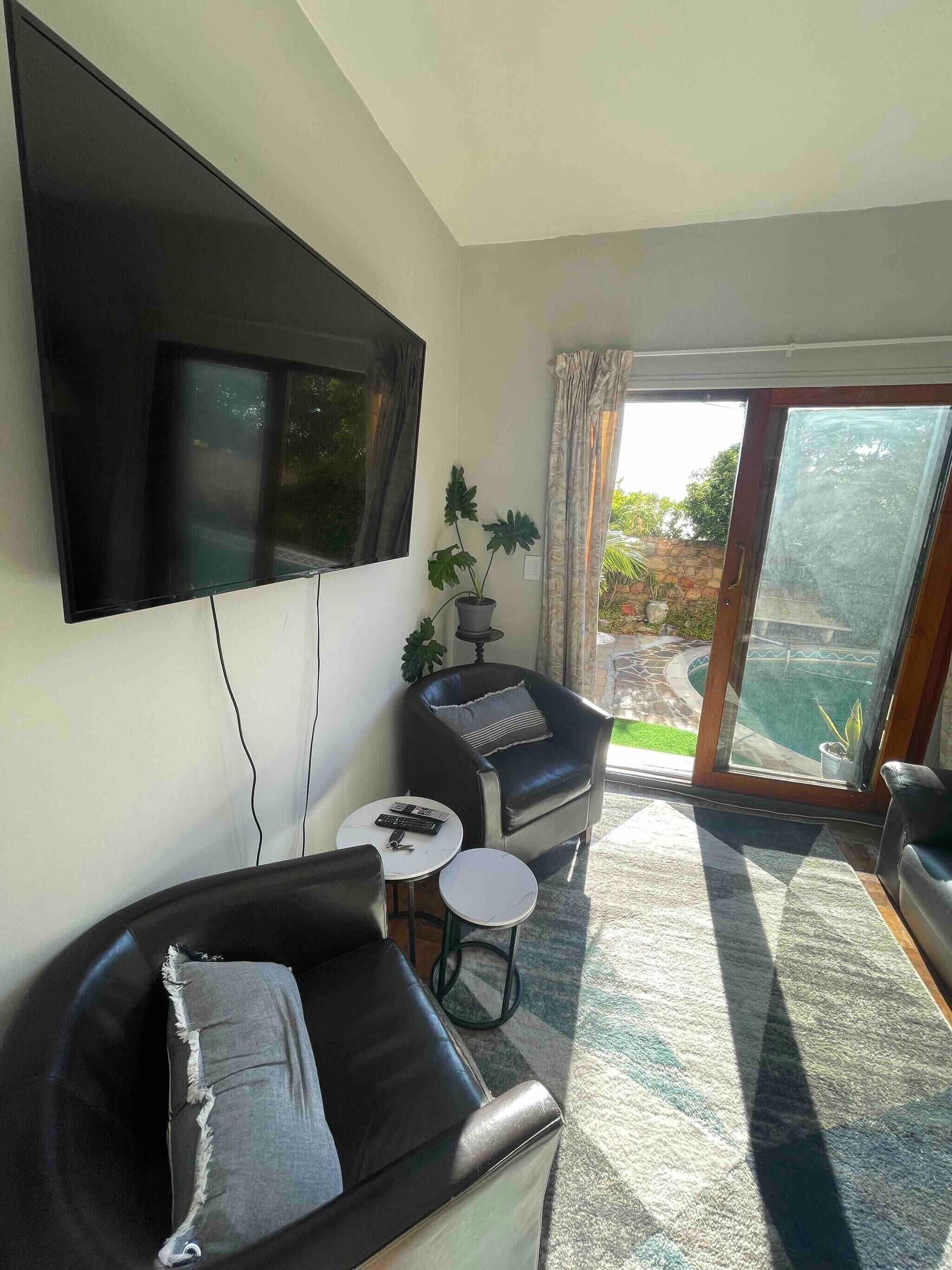 A living room with a black leather sofa, a small round table with a remote control, a potted plant, and a large TV mounted on the wall.
