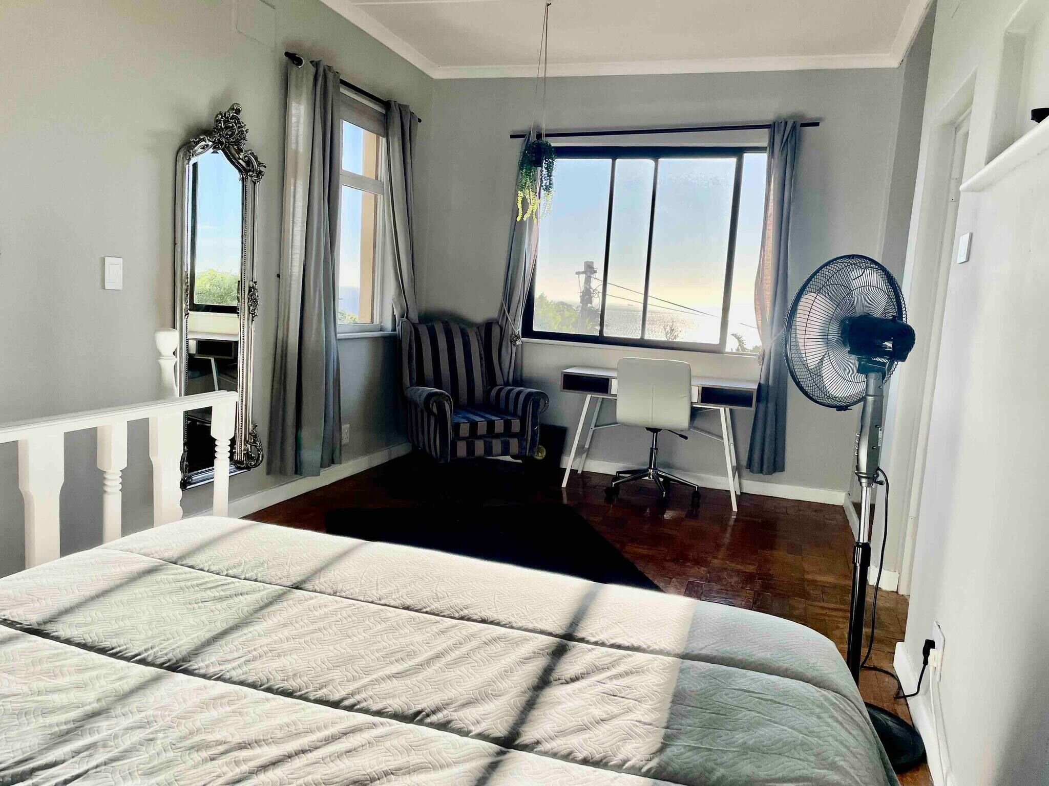 A bedroom with a bed, a desk, a chair, a standing fan, and a window with curtains.